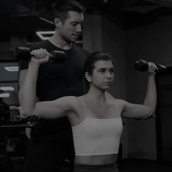 A coach standing behind a client, guiding a dumbbell shoulder press with both hands