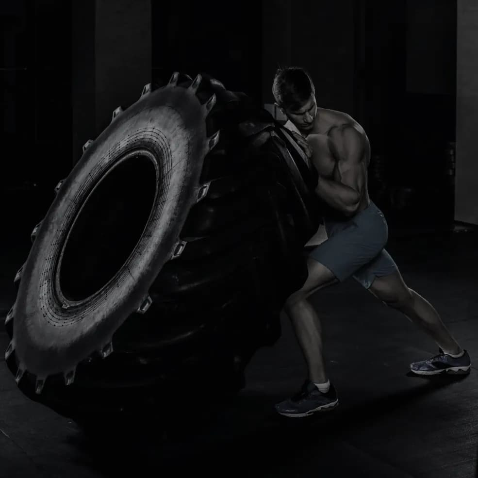 An athlete braced against a tractor tyre mid-flip on a gym floor