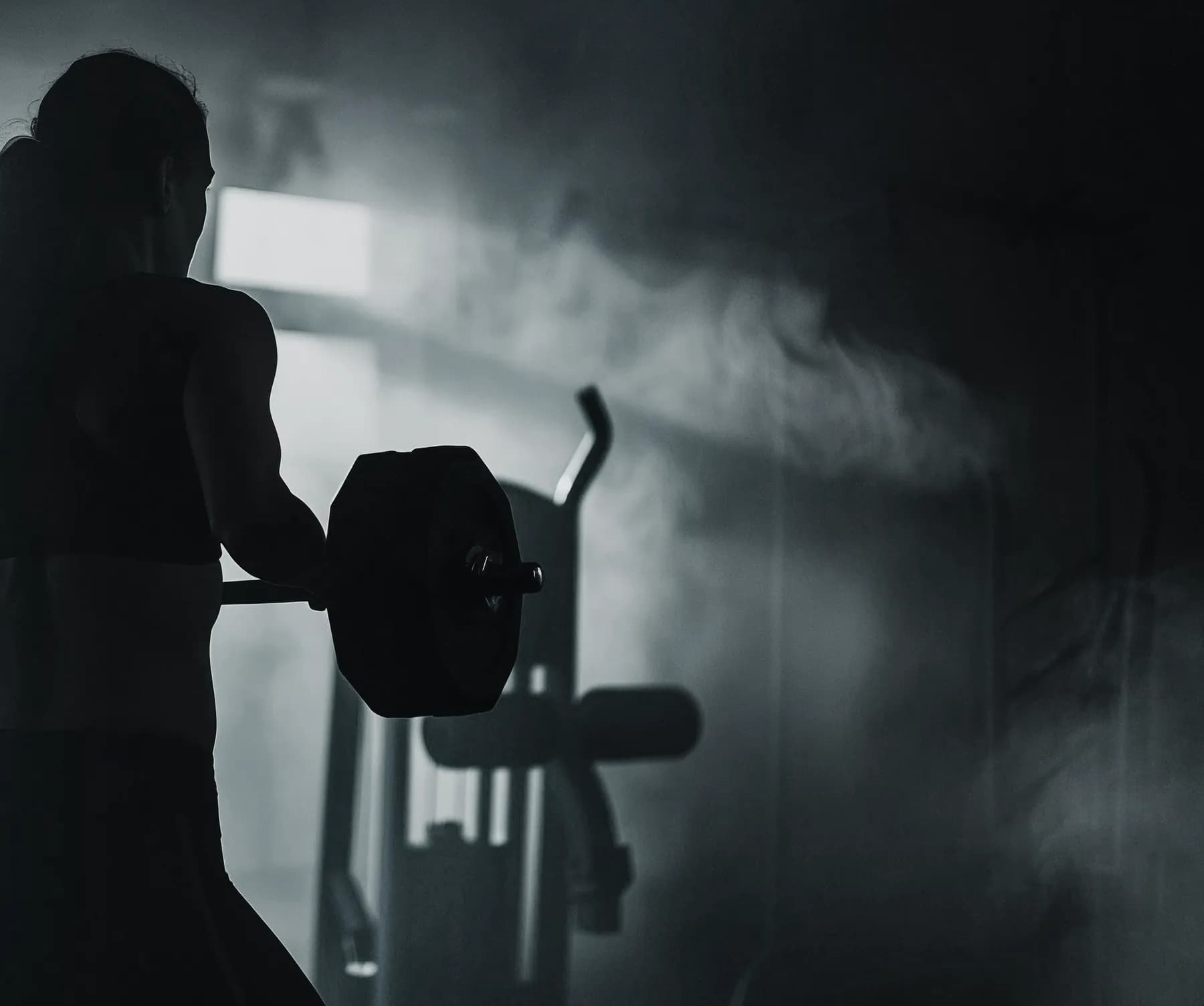 A lifter holding a loaded barbell at the hip, backlit through the smoke of a dark gym