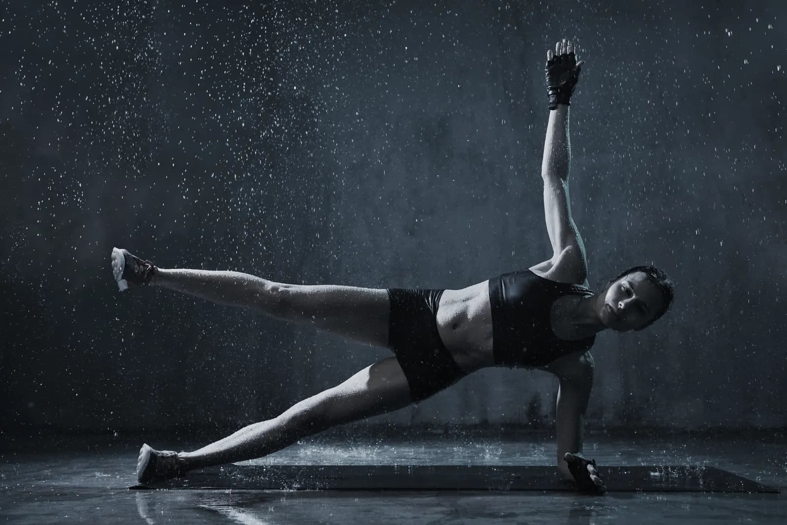 An athlete holding a side plank on a mat, arm raised, under falling water in a dark room